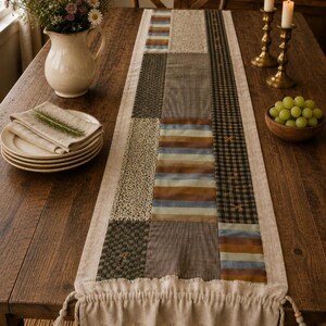 May include: A rectangular table runner featuring a patchwork design of various fabric patterns including stripes, checks, and solid textures in muted earth tones. The runner has a ruffled edge with decorative tassels at each corner of the ends.