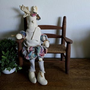 May include: A handmade doll with a deer-like head, wearing a cream-colored sweater, plaid skirt, and gray socks. The doll has cream-colored boots and is seated on a wooden chair with a woven seat. A small plant in a white pot is in the background.