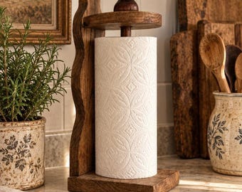 May include: A dark brown wooden paper towel holder with a vertical wavy side support, a flat rectangular base, and a rounded top finial. A roll of white paper towels with a repeating embossed geometric pattern is mounted on the central spindle.