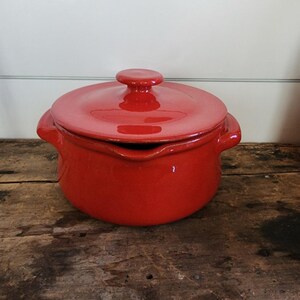 May include: A red ceramic casserole dish with a lid. The dish has two handles and a small spout. The lid has a round knob. The dish is sitting on a wooden surface.