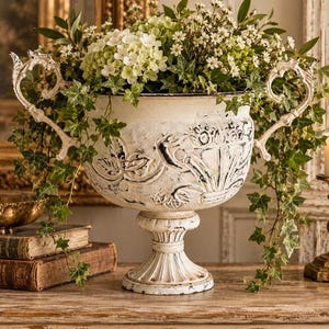 Puede incluir: Una maceta de metal blanco con acabado desgastado, que presenta asas ornamentadas en forma de voluta y patrones florales en relieve. El recipiente está lleno de un exuberante arreglo de hiedra verde, hortensias blancas y flores pequeñas.