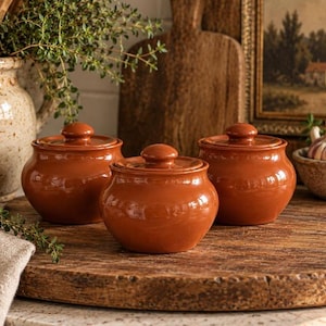 May include: Three matching terracotta pots with rounded bodies and fitted lids. Each pot features a smooth, glossy, reddish-brown finish, a short neck, and a small knob handle on the center of the lid. The pots are arranged in a cluster.