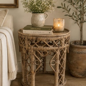May include: A round side table constructed from woven rattan with an intricate lattice pattern. The table surface holds a speckled ceramic pitcher filled with white flowers, two stacked books titled This Is Home and The Kinfolk Home, and a glass candle.
