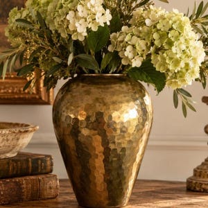 May include: A large, rounded, hammered brass vase with a metallic gold finish. The vase is filled with a lush arrangement of pale green and white hydrangea flowers and slender green leaves, creating a full, rounded floral display.