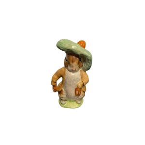 May include: A ceramic rabbit figurine wearing a green hat and a tan jacket. The rabbit is standing on a patch of green grass and holding a small orange object. The figurine has a detailed, hand-painted appearance.