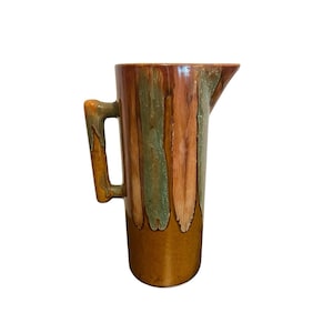 May include: A tall ceramic pitcher with a handle and spout. The pitcher has vertical stripes in brown, green, and tan. The base and handle are a solid tan colour. Likely used for serving beverages.