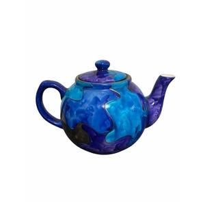 Puede incluir: Una tetera de cerámica con cuerpo redondeado, asa curva, boquilla y tapa extraíble. La superficie presenta un patrón jaspeado con tonos mezclados de azul profundo, turquesa brillante y morado oscuro, creando un efecto fluido y multicolor.
