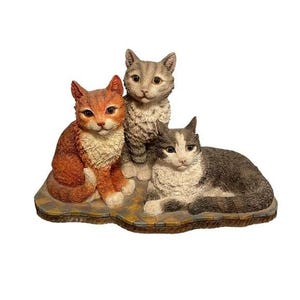 Puede incluir: Figura de cerámica con tres gatos. Uno es naranja, otro gris y el tercero gris y blanco. Están posados sobre una base a cuadros, con un gato sentado, otro de pie y otro reclinado.