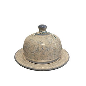 May include: A ceramic butter dish with a domed lid and matching plate. The dish is beige with blue speckles and a blue rim. A blue knob and a decorative string adorn the lid. The butter dish is designed to keep butter fresh.