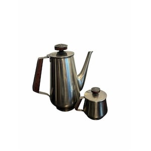 May include: A stainless steel coffee pot and a matching creamer set. Both pieces feature a brushed metallic finish, a tall tapered body, and dark brown wooden handles. Each lid is topped with a small, circular, textured wooden knob.