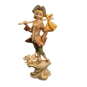 Puede incluir: Figura vintage de un joven con abrigo marrón, sombrero y pantalón verde, llevando un palo con un saco amarillo. Un perro pequeño está sobre una base decorativa. La figura tiene una estética clásica y antigua.