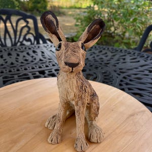May include: A sculpted hare figurine with a textured, light brown finish. The hare is depicted in a seated position with long, upright ears featuring dark brown tips, a dark nose, and black eyes. The surface of the sculpture shows detailed fur-like markings.