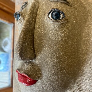 May include: A ceramic wall sculpture of a human face in profile. The sculpture has a textured, tan surface with dark blue eyes and red lips. The artwork is mounted on a wooden background.