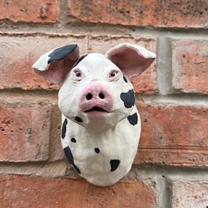 May include: A sculpted wall-mounted pig head figurine featuring a white base coat with irregular black spots. The pig has a prominent pink snout, dark eyes, and large ears with pink inner detailing, crafted with a textured, hand-modelled appearance.