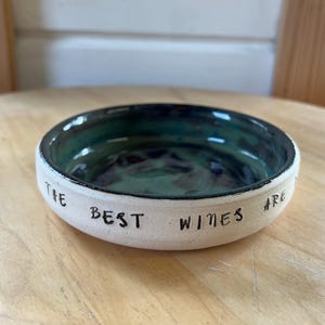 May include: A round ceramic bowl with a matte white exterior and a glossy, mottled teal and dark blue interior. The outer rim features the black stamped text "THE BEST WINES ARE". The bowl has a shallow, circular shape with a smooth, finished edge.