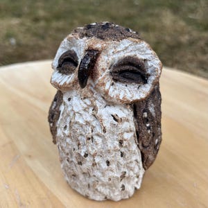May include: A small, hand-sculpted ceramic owl figurine featuring a textured, off-white body with dark brown speckled details. The owl has closed eyes, a dark brown beak, and a rough, matte finish across its entire surface, capturing a detailed avian form.