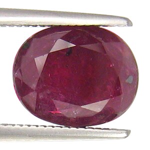 Puede incluir: Una piedra preciosa ovalada de color rojo oscuro con un corte facetado. La piedra preciosa está sujeta por unas pinzas.