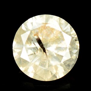 May include: A round, clear, faceted gemstone with a black inclusion in the center. The gemstone has a light yellow hue.