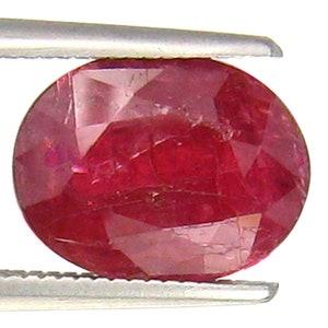 May include: A deep red oval-shaped gemstone with a faceted cut. The gemstone is held in a pair of tweezers.