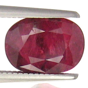 Puede incluir: Una piedra preciosa ovalada de color rojo oscuro con un corte facetado. La piedra preciosa se sostiene con unas pinzas.