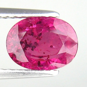 May include: A faceted, oval-shaped ruby gemstone held by silver tweezers. The ruby is a deep pinkish-red color with dark inclusions and internal fractures. The gem is cut to maximize light reflection.