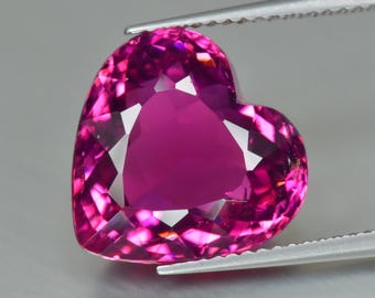 May include: A heart-shaped, faceted gemstone in shades of pink and magenta. The gem is held by silver tweezers, showcasing its polished surfaces and internal reflections. The gemstone's cut highlights its brilliance and clarity.