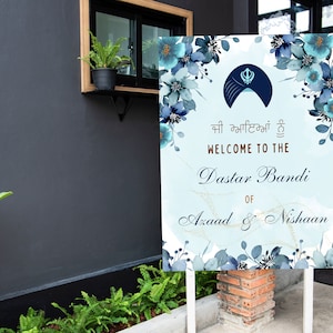 May include: A blue and white welcome sign with floral accents and gold glitter. The sign reads "Welcome to the Dastar Bandi of Azaad & Nishaan".