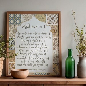 Könnte beinhalten: Gerahmtes Kunstwerk mit gemustertem Hintergrund und Kalligraphie auf einer weißen Tafel. Eine Holzschale, eine grüne Glasflasche und eine Vase mit Zweigen stehen auf einer Holzoberfläche. Ein religiöses Kunstwerk.