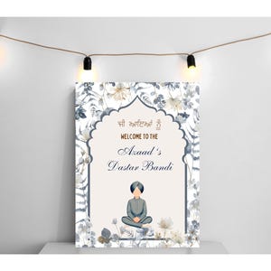 May include: A white sign with a floral border and a blue and white illustration of a person wearing a turban. The sign says "Welcome to the Azaad's Dastar Bandi" in gold lettering.