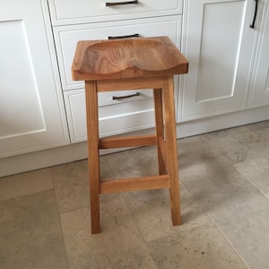 May include: A wooden bar stool with a curved seat and a simple, sturdy design. The stool is made of natural wood and has a light brown finish.