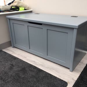 May include: A grey wooden storage bench with a hinged lid and a single pull-out drawer. The bench has a light grey finish and is sitting on a light wood floor.