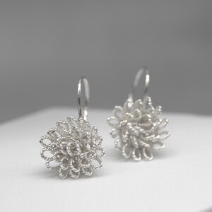 May include: A pair of silver-colored dangle earrings. Each earring features a floral design made of tightly wound wire. The earrings are displayed on a white surface against a soft gray background. The earrings have a hook closure.