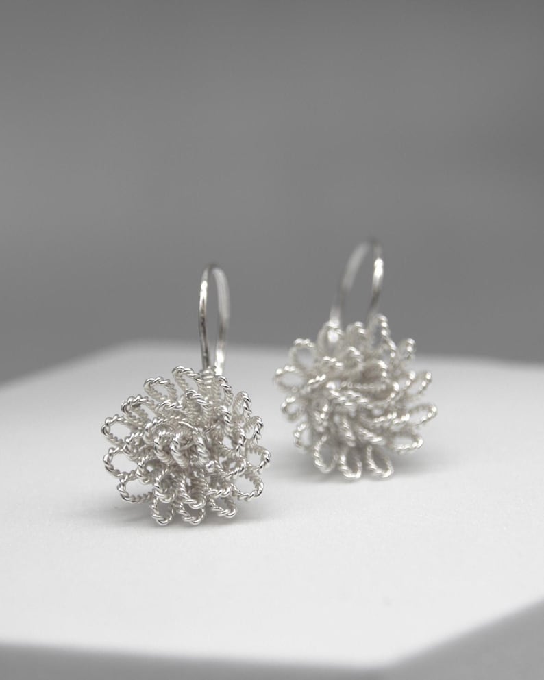 May include: A pair of silver-colored dangle earrings. Each earring features a floral design made of tightly wound wire. The earrings are displayed on a white surface against a soft gray background. The earrings have a hook closure.