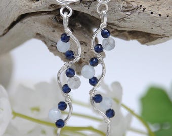 May include: A pair of silver wire-wrapped earrings featuring a spiral rose design at the top. Each earring has a dangling, wavy silver wire stem adorned with alternating dark blue faceted beads and pale, translucent light blue round beads.