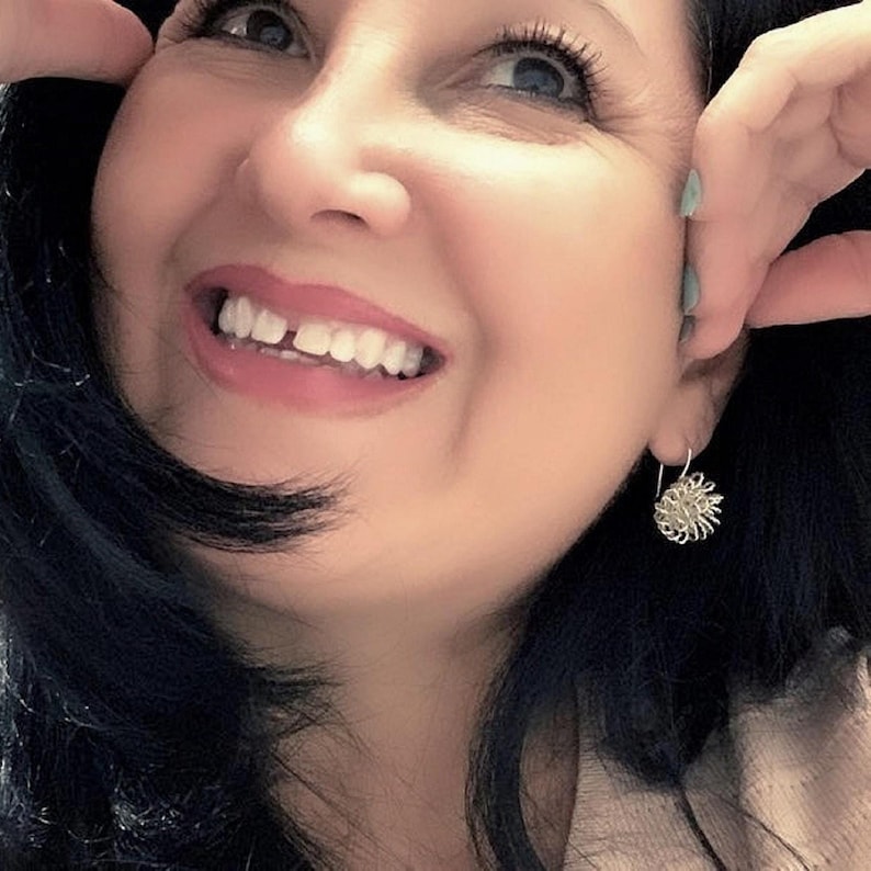 May include: A smiling person with dark hair and a light-colored top. The person is wearing a silver earring with a floral design. The person is looking up and to the right.