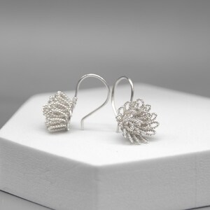 May include: A pair of silver-toned earrings. Each earring features a looped wire design, creating a textured, floral-like shape. The earrings are displayed on a white surface against a soft gray background.