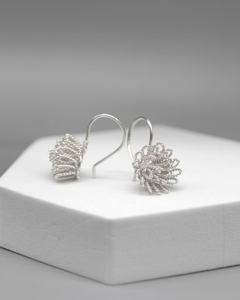 May include: A pair of silver-toned earrings. Each earring features a looped wire design, creating a textured, floral-like shape. The earrings are displayed on a white surface against a soft gray background.