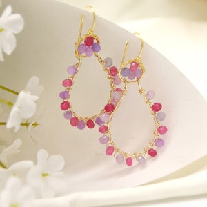 Può includere: Un paio di orecchini in filo d'oro con perline di pietre preziose rosa e viola. Gli orecchini hanno la forma di lacrime e pendono.