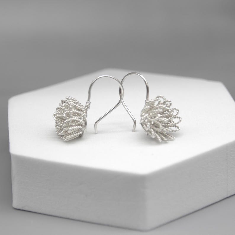 May include: A pair of silver-colored earrings. Each earring features a textured, woven design in a rounded shape, suspended from a simple hook. The earrings are displayed on a white surface against a soft gray background.