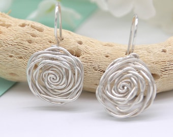 May include: A pair of silver-colored wire earrings. Each earring features a spiral design within a square frame, suspended from a simple hook. The earrings are displayed against a natural, textured background with white flowers.
