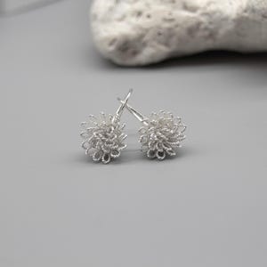 May include: A pair of silver-colored, wire-wrapped earrings. Each earring is a small, round cluster with a delicate, open design. The earrings are displayed on a light gray surface, with a textured, off-white stone in the background.