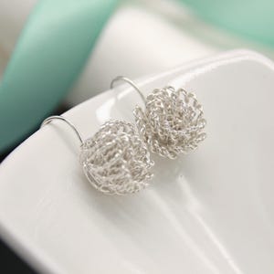 May include: A pair of silver-colored earrings. Each earring features a woven, spherical design suspended from a curved ear wire. The earrings are displayed on a white surface, with a soft, blurred background.