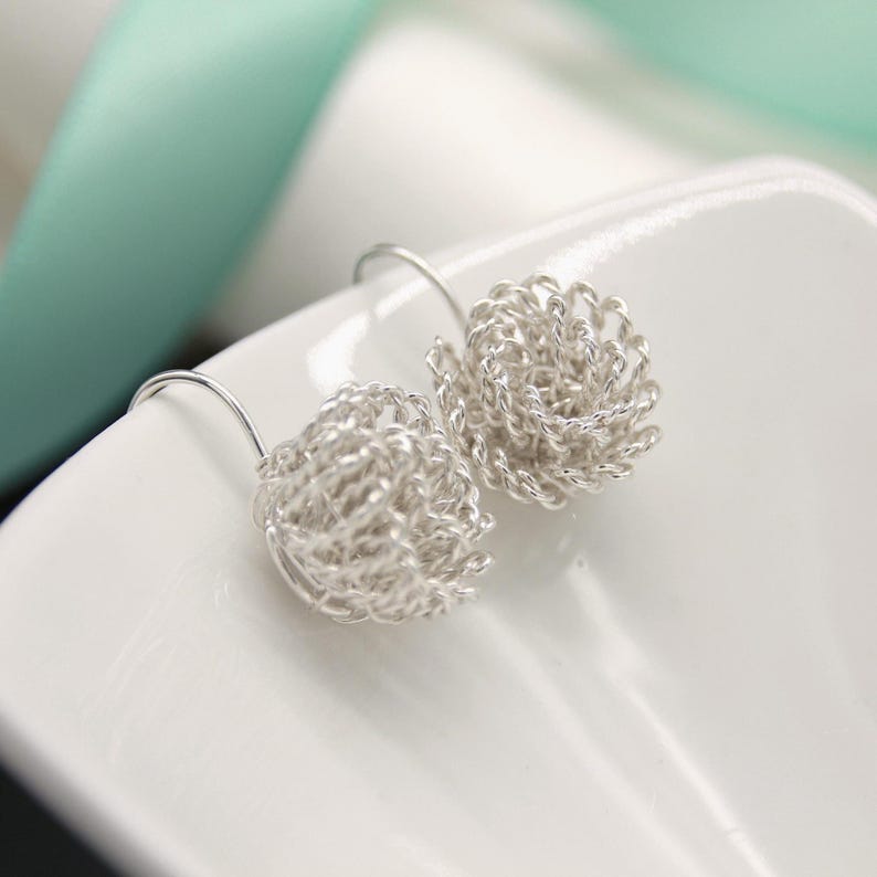 May include: A pair of silver-colored earrings. Each earring features a woven, spherical design suspended from a curved ear wire. The earrings are displayed on a white surface, with a soft, blurred background.