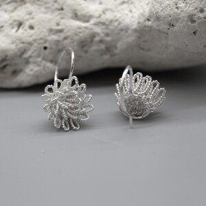 May include: A pair of silver-colored earrings. Each earring features a floral design made of woven wire. The earrings have a hook closure and are displayed on a gray surface with a textured rock in the background.