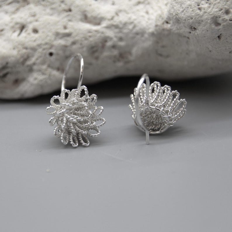 May include: A pair of silver-colored earrings. Each earring features a floral design made of woven wire. The earrings have a hook closure and are displayed on a gray surface with a textured rock in the background.