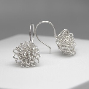May include: A pair of silver-colored earrings. Each earring features a floral design made of tightly woven wire. The earrings have a hook closure. The background is a soft, neutral gray and white.