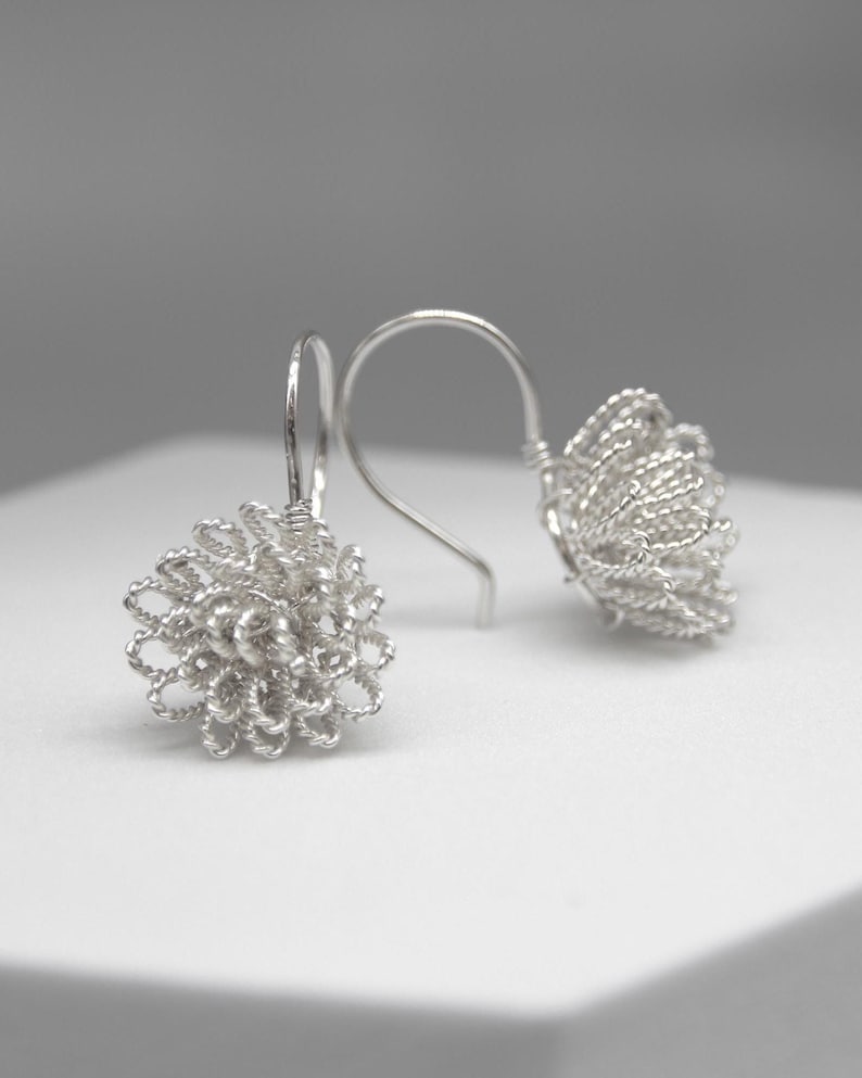 May include: A pair of silver-colored earrings. Each earring features a floral design made of tightly woven wire. The earrings have a hook closure. The background is a soft, neutral gray and white.