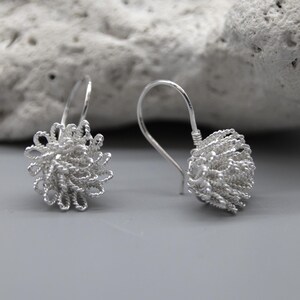 May include: A pair of silver-colored earrings. Each earring features a looped wire design forming a spherical shape, suspended from a curved ear wire. The earrings are displayed against a neutral gray background with a textured white stone.