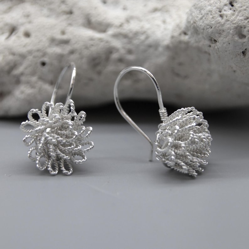 May include: A pair of silver-colored earrings. Each earring features a looped wire design forming a spherical shape, suspended from a curved ear wire. The earrings are displayed against a neutral gray background with a textured white stone.