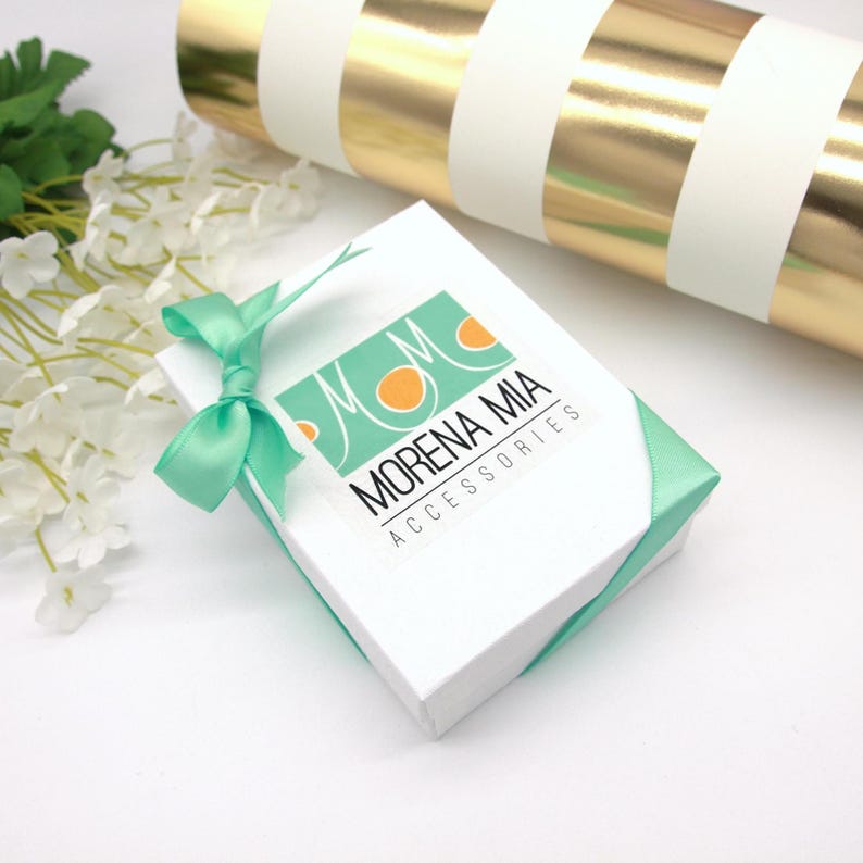 May include: A white square gift box with a teal ribbon and bow. The box has the words "MORENA MIA ACCESSORIES" printed on it. A roll of gold and white striped wrapping paper is in the background.